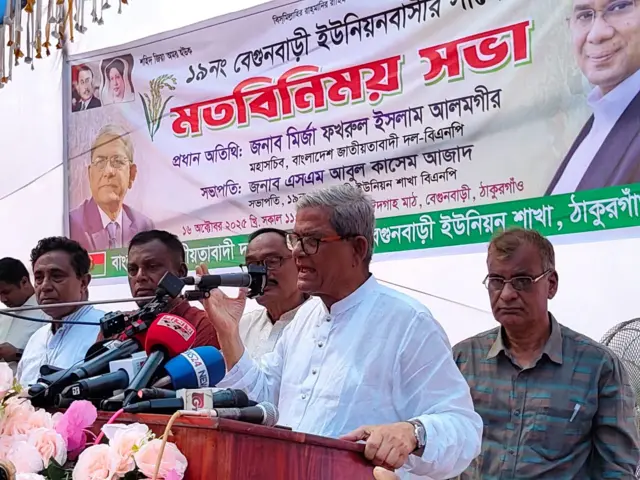 মির্জা ফখরুল ইসলাম আলমগীর দাঁড়িয়ে টেবিলে হাত রেখে মাইকের সামনে কথা বলছেন, পাশে দাঁড়িয়ে আছে কয়েকজন