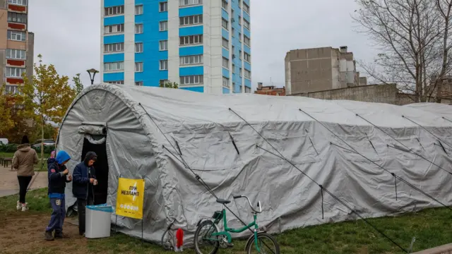 Серая цилиндрическая палатка стоит среди многоэтажных домов, на входе вывеска "пункт незламностi", у входа стоит велосипед