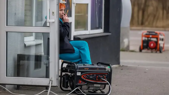 Женщина в верхней одежде сидит у генератора перед сооружением из стеклопакетов