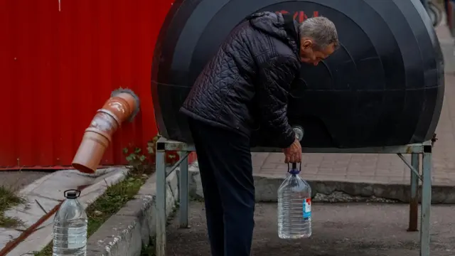 Мужчина в болоньевой куртке наливает воду в пластиковую бутыль из цистерны
