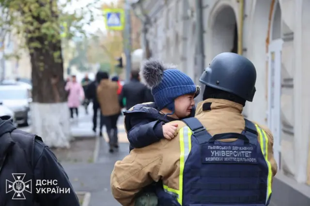 Сфотографированный со спины мужчина в синем жилете с надписью "Львовский университет безопасности жизнедеятельности" несет на руках плачущего мальчика в шапке с помпоном