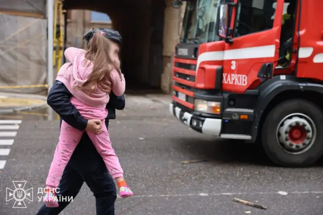 Мужчина несет на руках девочку в розовой одежде, рядом стоит машина с надписью "Харьков" на украинском языке