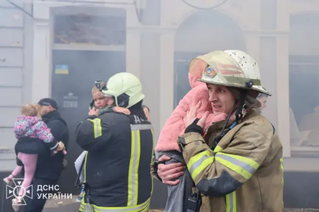 Мужчины в защитных костюмах и касках со светоотражающими полосами несут на руках маленьких детей