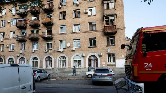 Многоэтажный кирпичный дом с выбитыми окнами и поврежденными балконами, с краю виден грузовик с надписью "Киев" на украинском языке