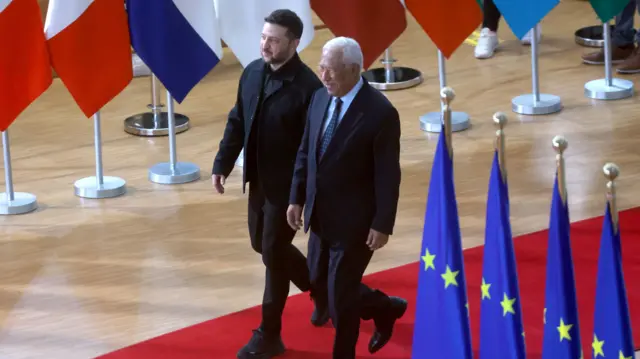 Зеленский в темном костюме и рубашке идет по красной ковровой дорожке вместе с Коштой в темном костюме и светло-синей рубашке. На переднем плане видны флаги ЕС