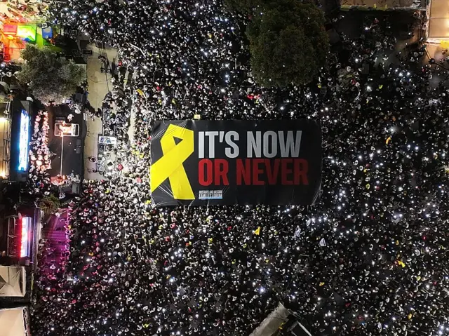 تظهر الصورة تجمع إسرائيليين من الأعلى ليلاً، في احتجاج مطالب باستعادة الرهائن، يحملون يافطة كبيرة لونها أسود كتب عليها باللغة الإنجليزية "it's now or never" بجانبها الإشارة الصفراء لدعم الرهائن.
