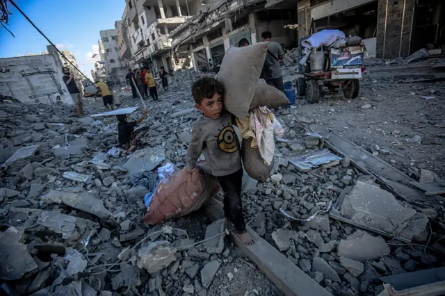 طفل فلسطيني يحمل أمتعة على ظهره ويمشي حافي القدمين على أنقاض مبانٍ مدمرة في غزة.