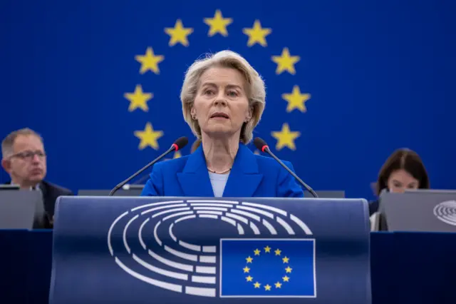 Урсула фон дел Ляйен во время выступления в Европарламенте. Она стоит на трибуне с флагом Евросоюза, ее голову окружают желтые звезды из флага ЕС