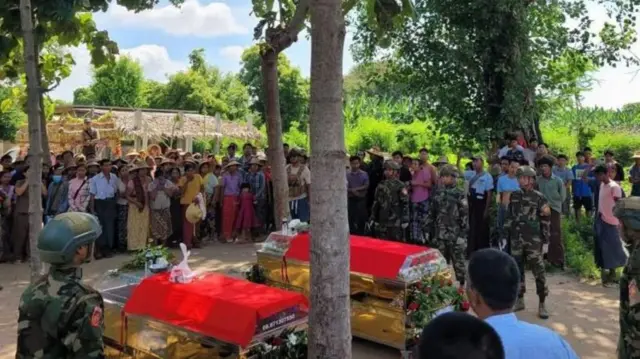 দুটি কফিনের পাশে স্থানীয় লোকজন ও মিলিশিয়া সদস্যরা দাঁড়িয়ে আছেন