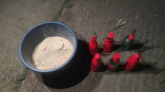 লাল স্কচটেপে মোড়ানো ছয়টি পেট্রলবোমা রাখা, একটি বালতির ভেতরে বালুর মধ্যে রাখা হয়েকটি হাতবোমা