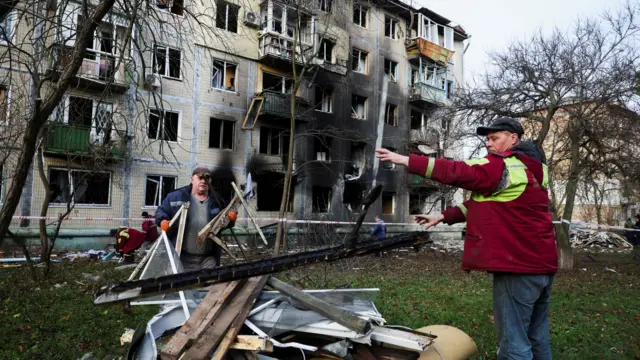 Пятиэтажный дом, на уровне 2-3 этажей видны последствия пожара. На переднем плане двое мужчин разбирают обломки.