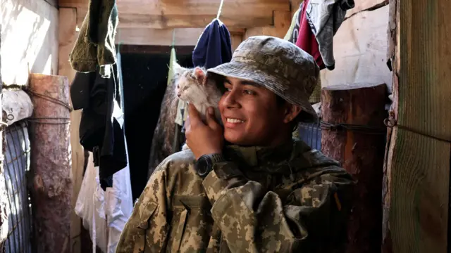 Смуглый юноша в камуфляже с белым котенком на плече в блиндаже