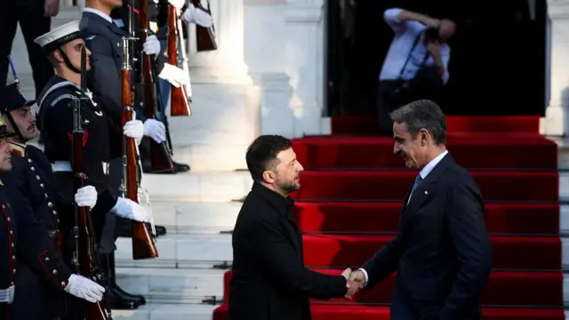 Владимир Зеленский и Кириакос Мицотакс пожимают друг другу руки у лестницы, покрытой красным ковром, слева строй солдат почетного караула, на заднем плане человек с фотокамерой