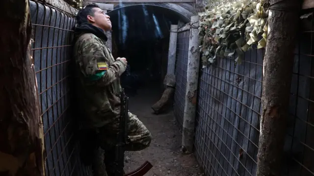 Мужчина в камуфляже с нашивкой в виде флага Колумбии на рукаве курит в подземном укрытии, прислонив автомат к стене