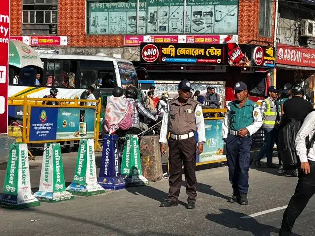 নতুন ও পুরোনো পোশাকে দুই জন ট্রাফিক পুলিশ দাঁড়িয়ে আছেন, পেছনে কয়েকটি যানবাহন দেখা যাচ্ছে