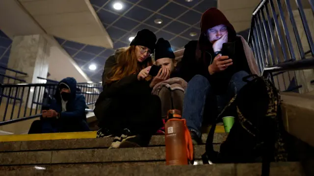 Мужчина, женщина, ребенок и поодаль еще один мужчина сидят на ступеньках лестницы, смотрят в телефон и кутаются в верхнюю одежду