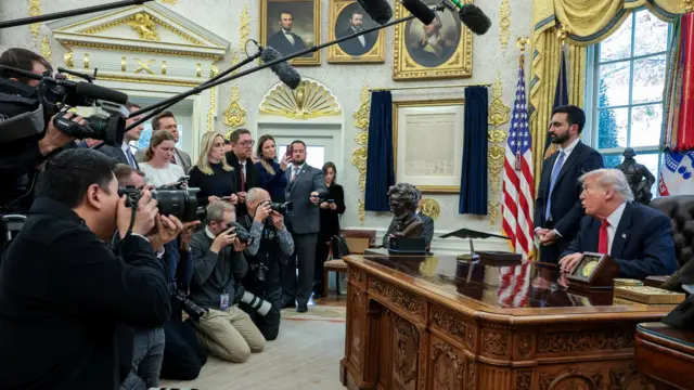 Толпа репортеров с камерами и микрофонами перед Дональдом Трампом, который сидит за столом Resolute, и Зохраном Мамдани, который стоит рядом с ним