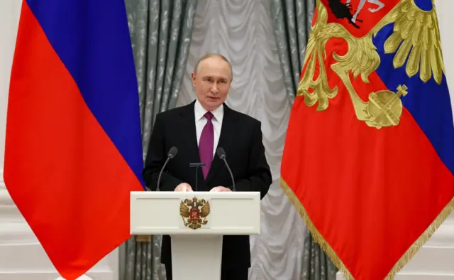 Владимир Путин стоит у трибуны с двумя микрофонами и двухглавым орлом, у него за спиной два российских флага, один из них с гербом