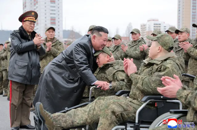 Ким Чен Ын в кожаном плаще стоит и обнимает мужчины в военной форме на инвалидном кресле.
