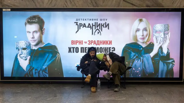 Два человека дремлят на стульях под рекламой телешоу "Зрадники"