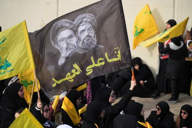سيدة تحمل صورة القادة الراحلين لحزب الله حسن نصر الله وهاشم صفي الدين، وحولها عدد من النساء يرتدون لباساً أسود ويحملون رايات الحزب الصفراء.