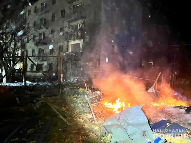 Поврежденная при ударе многоэтажка, рядом на земле что-то горит.