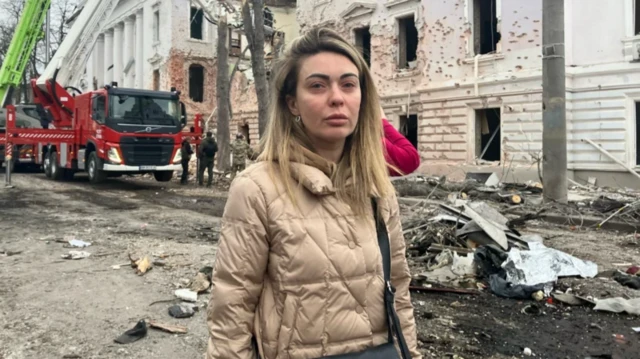 Молодая женщина в куртке, на заднем плане здание с выбитыми стеклами в окнах и пожарные машины