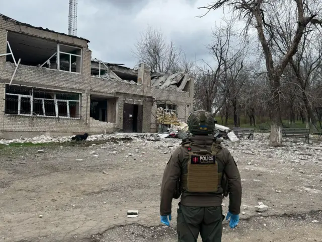Человек в бронежилете с надписью Police осматривает двухэтажное здание, поврежденное в результате обстрела.