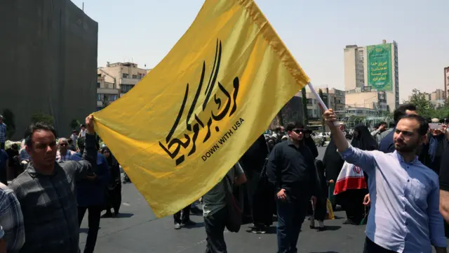 إيرانيون يحملون علماً كُتب عليه "تسقط أمريكا" خلال احتجاج ضد الغارات الجوية الإسرائيلية في طهران.