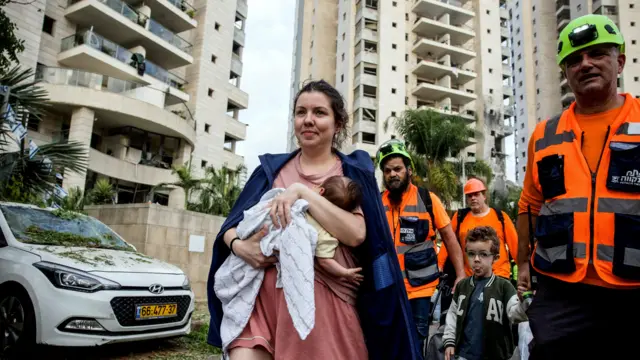 امرأة تحمل طفلها بمرافقة فرق الإنقاذ أثناء مغادرة مجمع سكني متضرر في أعقاب هجوم صاروخي من إيران على إسرائيل.