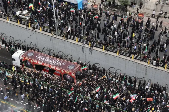 موكب يظهر فيه أحد النعوش ويعتليه نص من آية قرآنية "نصر من الله وفتح قريب". حملت الجموع الأعلام الإيرانية