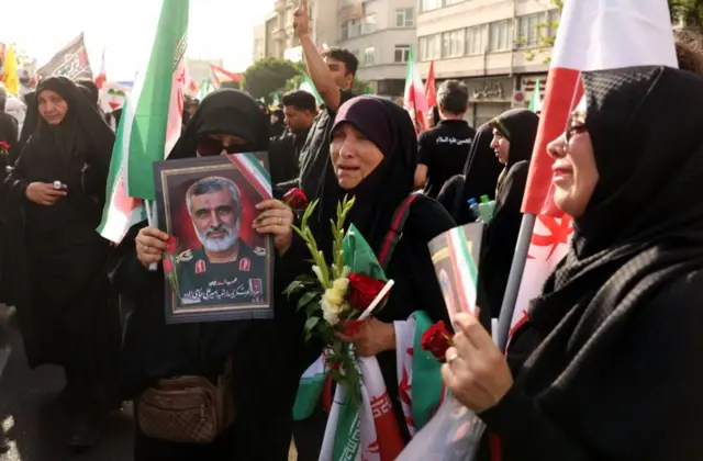 نساء يرتدين الأسود ويبكين، وتحمل إحداهن صورة الراحل أمير علي حاجي زاده، قائد القوة الجوفضائية التابعة للحرس الثوري الإيراني، وأخريات حملن أعلام دولة إيران وخلفهم مجموعة من الرجال بلباس أسود أيضاً.