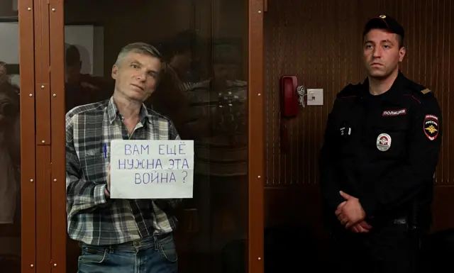 Алексей Горинов в стеклянной клетке показывает плакат: ВАМ ЕЩЕ НУЖНА ЭТА ВОЙНА?