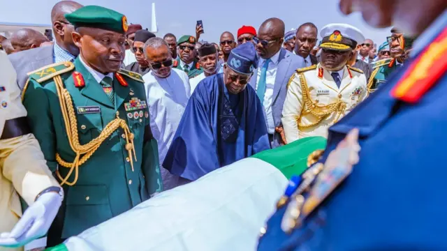 A military official looking at di remains of Buhari covered wit Nigeria flag