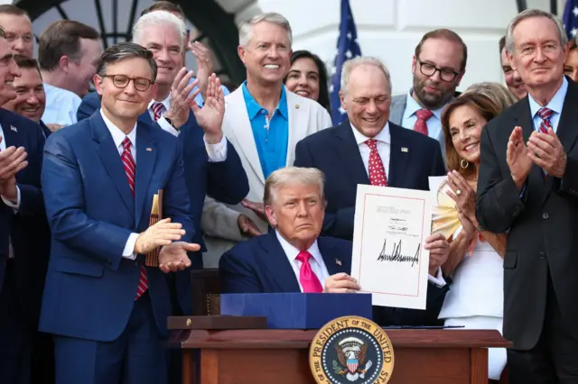 Onyeisiala Trump nọ ọdụ n'oche dịka ndị ọrụ ya gbara ya gburu gburu na-akụrụ ya aka.