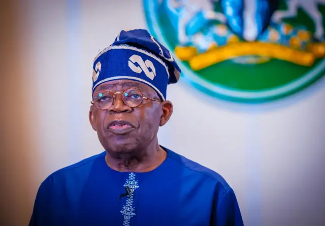 Bola Tinubu sanye da shuɗin tufafi da shuɗiyar riga, tsaye sanye da tubarau, yayin da aka maƙala abin ɗaukar magana a rigarsa