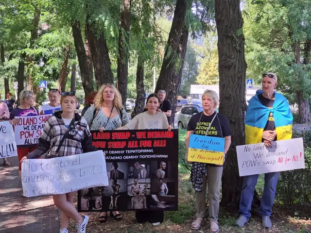 Близько десяти людей стоять на алеї біля дороги, в руках тримають плакати, на яких англійською мовою написані звернення