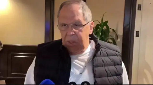 Сергій Лавров у приміщенні, схожому на лоббі, у білому светрі та чорній жилетці