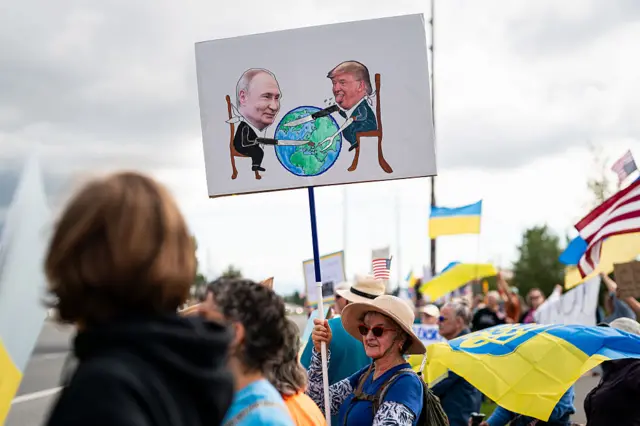 Літня американка тримає над головою плакат, на якому символіячний малюнок-колаж: Путін і Трамп, сидячи за Землею (планетою), як за столом, у "слюнявчиках", ріжуть планету, а Путін всаджує ніж в Україну. На задньому плані десятки людей з українськими та американськими прапорами