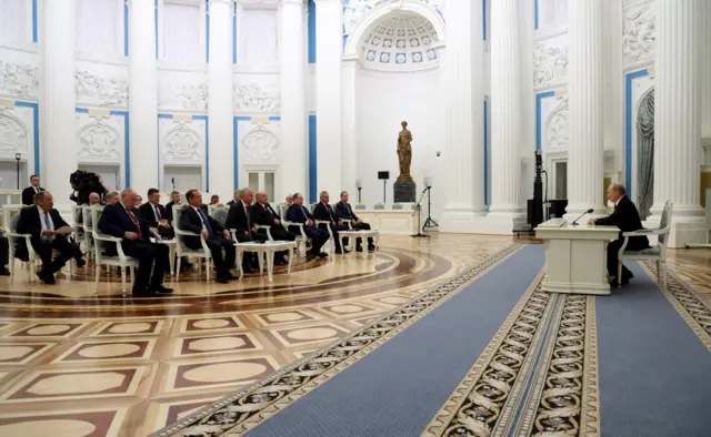 Путин сидит в кресле за столом на большом расстоянии от нескольких рядов стульев с официальными лицами
