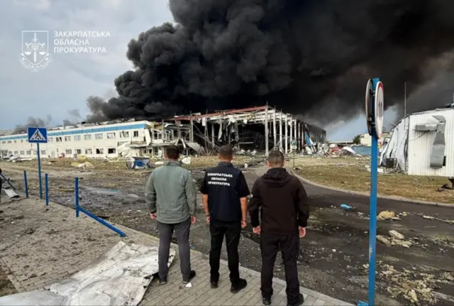 Три человека смотрят на развалины двухэтажного склада, разрушенного в результате удара. Над зданием поднимается облако дыма