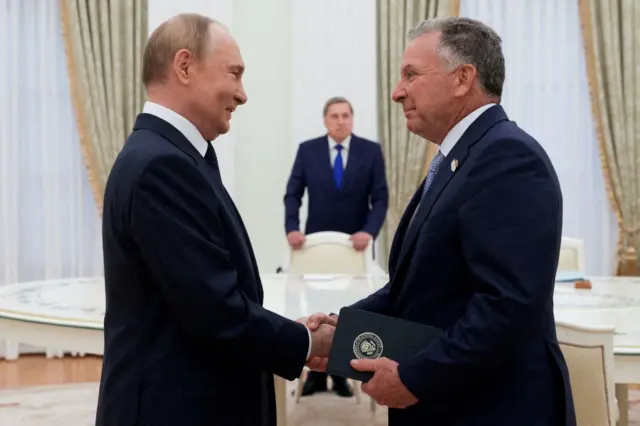 Владимир Путин и Стивен Уиткофф жмут друг другу руки во время встречи в Кремле. За ними стоит человек в синем костюме и галстуке