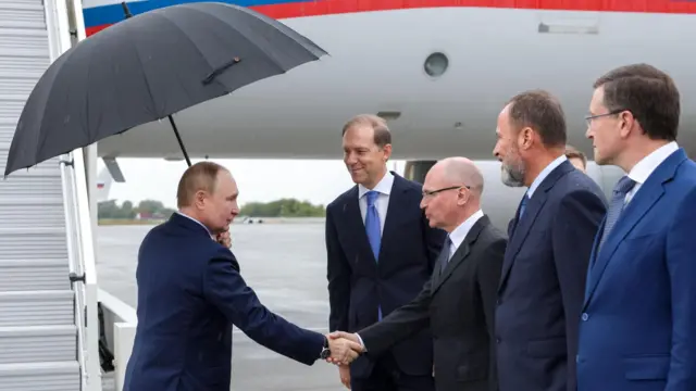 Путин одной рукой держит зонтик у трапа самолета, другой поживает руку Сергею Кириенко на летном поле. Рядом с Кириенко еще три чиновника высокого ранга улыбаются