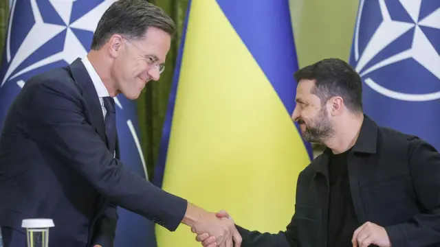 Марк Рютте и Владимир Зеленский жмут друг другу руки, стоя на трибуне на фоне флагов Украины и НАТО