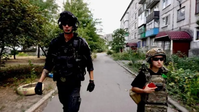 Двое людей — высокий мужчина в черной одежде и невысокая женщина в камуфляже цвета хаки, оба в бронежилетах и темных очках — идут на фоне многоэтажных домов.