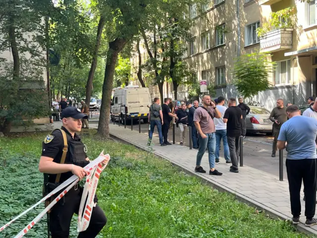 Полицейский оцепляет место преступления, люди стоят рядом.