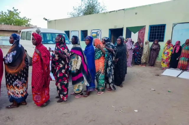 Wata mata sanya ta tufafi masu launi daban-daban a kan layin karɓar tallafi a gaban wani gini