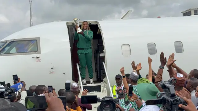 Kaftin ɗin tawagar Najeriya riƙe da kofi yayin da take fitowa daga jirgi mutane na murna a bakin jirgin