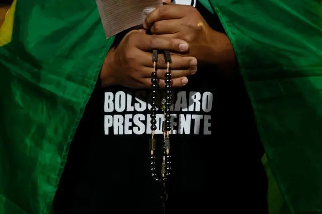 Mão de mulher com um terço rezando, com camiseta escrita Bolsonaro Presidente