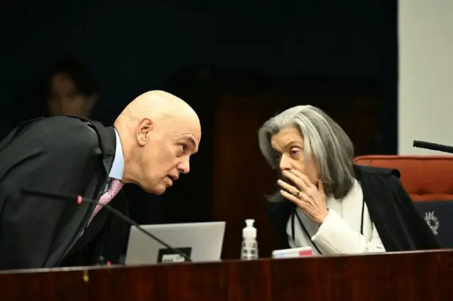 Moraes e Cármen Lucia cochichando; ele está em pé, se aproximando dela, que está sentada